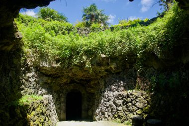 Hallim Park 'taki lav mağarasına açılan kemerli bir kapı üzerinde yemyeşil bitki örtüsüyle çevrili obruk benzeri bir çatı penceresi açılıyor..