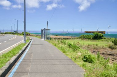 Jeju Şehri, Güney Kore - 1 Temmuz 2025: Bir cam otobüs durağı ve sahil patikası Sinchang yakınlarındaki Iljuseo Yolu 'nu takip ediyor, dalgakıranların ve ağların ötesinde dönen bir dizi rüzgar türbini var.