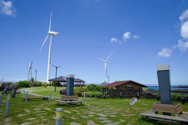 Jeju Şehri, Güney Kore - 1 Temmuz 2025: Gazebos ve küçük bir park Sinchang Yel Değirmeni Sahil Yolu 'ndaki uzun rüzgar türbinlerinin altında asfalt yollar, heykeller ve denize bakan manzaralı..