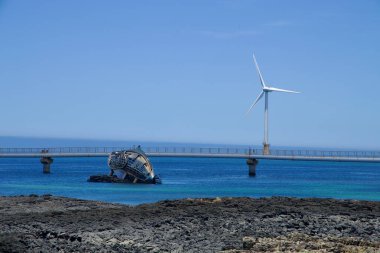 Jeju Şehri, Güney Kore - 1 Temmuz 2025: Metal bir gemi enkazı kıyı kayaları üzerinde dinlenirken, açık deniz rüzgar türbini ve yaya köprüsü Sinchang Yel Değirmeni Sahil Yolu 'ndaki mavi suyu kaplıyor.