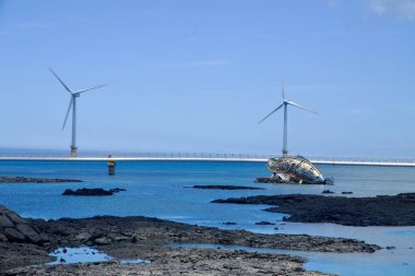 Jeju Şehri, Güney Kore - 1 Temmuz 2025 Eğik gemi enkazı heykeli Sinchang Yel Değirmeni Sahil Yolu 'nun kenarında, körfez boyunca ikiz açık deniz rüzgar türbinleriyle duruyor..