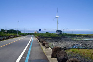 Jeju Şehri, Güney Kore - 1 Temmuz 2025 Sinchang Yel değirmeni Sahil Yolu yakınlarındaki bir rüzgar türbinine doğru kıyı yolu ve mavi bisiklet yolu kavisi, taş duvarlar ve gelgit düzlüklerinden geçiyor..