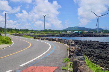Jeju Şehri, Güney Kore - 1 Temmuz 2025: Sahil yolu ve adanmış mavi bisiklet yolu Sinchang Windmill Sahil Yolu 'ndaki bazalt kayaları ve rüzgar türbinlerini geçerek iç kısımlara doğru yükselen orman tepeleriyle birlikte.
