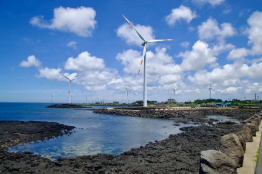 Jeju Şehri, Güney Kore - 1 Temmuz 2025 Sinchang Yel Değirmeni Sahil Yolu 'nda dalgaların ve kümülüs bulutlarının sürüklendiği karanlık volkanik kayaların olduğu kıyıya çok sayıda rüzgar türbini dağıldı..