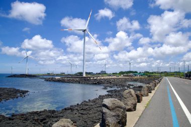Jeju Şehri, Güney Kore - 1 Temmuz 2025: Bir sıra rüzgâr türbini Sinchang Yel değirmeni Sahil Yolu 'ndaki mavi bisiklet yolunun kıyısında ön planda bazalt çıkıntıları ve durgun suları olan bir sahil şeridi oluşturur..