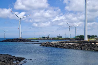 Jeju Şehri, Güney Kore - 1 Temmuz 2025: Kayalık burunların açık denizle buluştuğu Sinchang Windmill Sahil Yolu 'ndaki evlerin ve deniz kenarındaki rüzgar türbinlerinin yakın görüntüsü.
