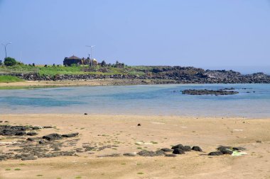 Sindo Limanı yakınlarındaki sakin bir koy, Jeju 'nun batı kıyısındaki körfezi çevreleyen, soluk kum, temiz sığ ve dağınık lav kayaları, küçük bir pavyon ve alçak yeşil bir set göstermektedir..