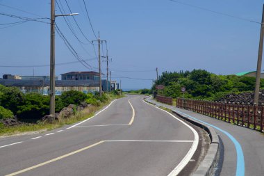 Seogwipo City, Güney Kore - 3 Temmuz 2025: Çitli bir bisiklet yolu ile kıvrımlı bir kıyı yolu Onpyeong 'a doğru ilerliyor, elektrik direkleri olarak atölyeleri ve çalıları geçiyor.