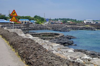 Seogwipo Şehri, Güney Kore - 3 Temmuz 2025: Dairesel taş havuzlar ve lav deniz duvarları Onpyeong kıyı şeridi, mavi sığ ve siyah kaya çıkıntıları üzerindeki köy işaretleri.