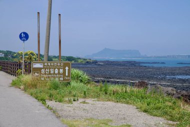 Seogwipo Şehri, Güney Kore - 3 Temmuz 2025: Bisiklet ve yaya yolu üzerindeki bir kıyı işareti Seongsan Ilchulbong 'u işaret ediyor, volkanik koni lav düzlüklerinin ve gelgit havuzlarının ötesinde görülebilir..