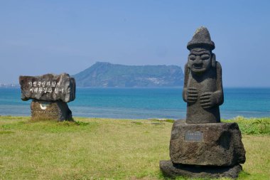 Seogwipo Şehri, Güney Kore - 3 Temmuz 2025: Seopjikoji kıyısında bir dol hareubang heykeli bulunuyor, Seongsan Ilchulbong 'un ötesinde bir kayayla eşleştiriliyor..