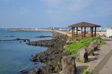Ahşap pavyonu olan bir sahil yürüyüşü Seongsan 'daki limana doğru bazalt kayalarını takip eder. Sarı dalgakıran deniz fenerleri ve feribotları görülebilir..