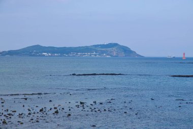 Udo Adası, Dumun Limanı 'nın ötesindeki ufuk boyunca uzanır ve ön planda katmanlı bazalt çıkıntıları ve durgun su ile kanal girişini gösteren parlak turuncu bir deniz feneri oluşturur..