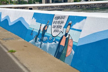 Jeju Haenyeo dalgıçlarının renkli bir duvar resmi bisiklet yolu boyunca Handong Denizi 'ni süsler ve kayalık sahilde sepetleri olan çizgi film yüzücülerini gösterir..