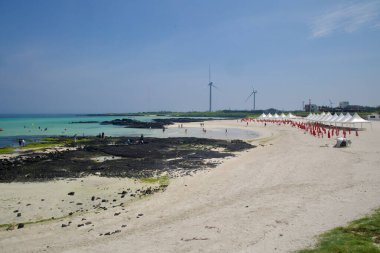 Gimnyeong Seongsegi Sahili 'nin geniş bir görüntüsünde turkuaz sığlıklar, bazalt çıkıntıları ve kırmızı şemsiyeli beyaz örtüler ve ufukta rüzgar türbinleri görülüyor..