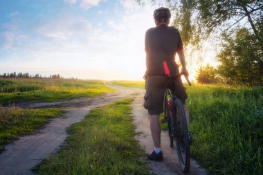 Gün batımında yolda bisiklet süren bir adam. Bisikletçi, bisiklet, arazi, yeşil çimenler, ağaçlar ve parlak güneş ışığıyla renkli bir manzara. Spor ve seyahat.