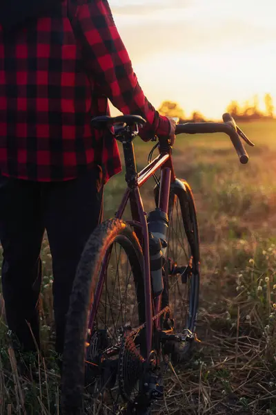 Close-up shot of active cyclist wearing a checkered shirt, holding a gravel bike in a field against a stunning golden sunset. Active lifestyle, sports, and leisure.
