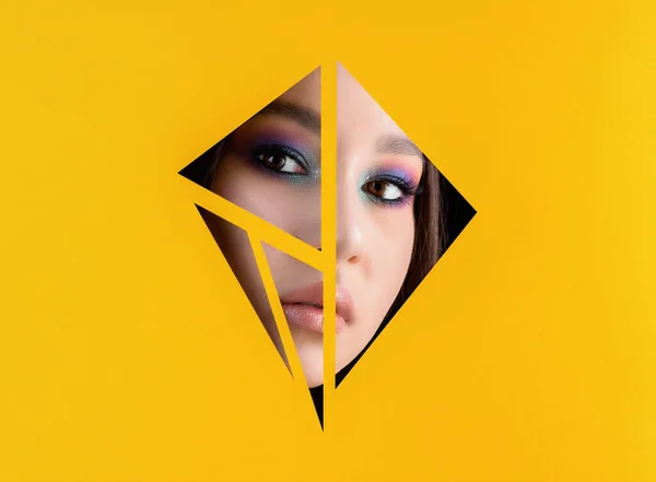 Beautiful young girl with make up looking through triangles in yellow paper background. Space for text.