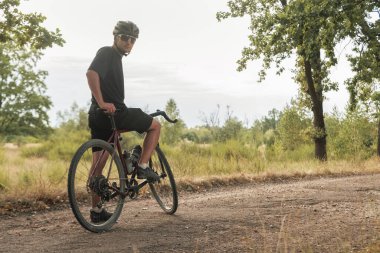Gözlüklü ve kasklı genç bir bisikletçi kırsaldaki çakıllı bir yolda bisikletle duruyor..