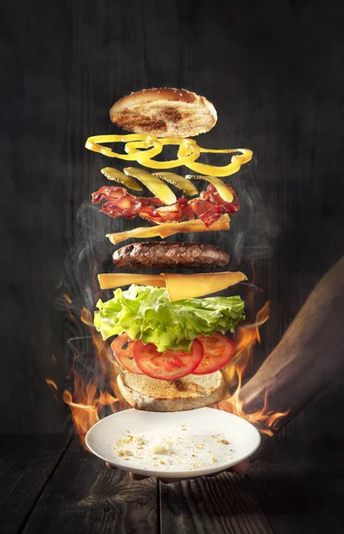 Tasty hamburger with flying ingredients. A hamburger flies over a plate with fire and smoke. Wooden background.