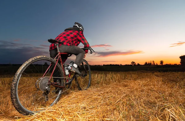 A cyclist on a gravel bike stands in a field against the backdrop of the sunset. Low angle view.