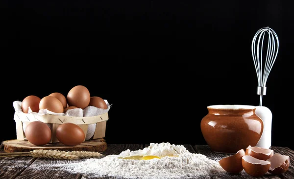 Frozen photo of a broken egg falling in flour. Flour is poured on a wooden board with a texture. In the background is a box with eggs, a whisk and a pot. Dough preparation.