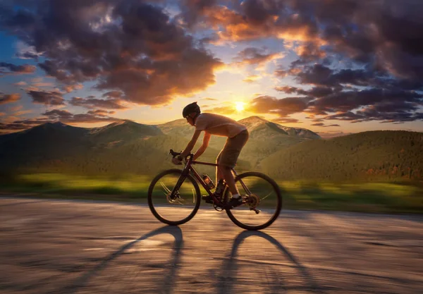 Cyclist riding on a mountain road at sunset. Beautiful landscape of man on bicycle riding on asphalt road at beautiful mountains background with dramatic sunset sky. Soft focus.