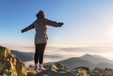 Yükselmiş elleri ile genç mutlu bir kadın alçak bulutlarda bir dağın zirvesinde güneşin doğuşunu seyrediyor..
