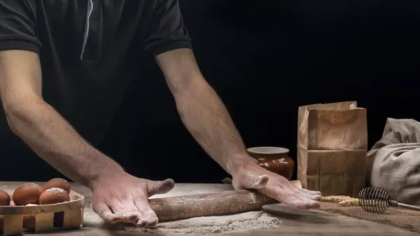 Man cooks homemade bread in the kitchen. Baker rolls dough with a rolling pin on a wooden table sprinkled with flour