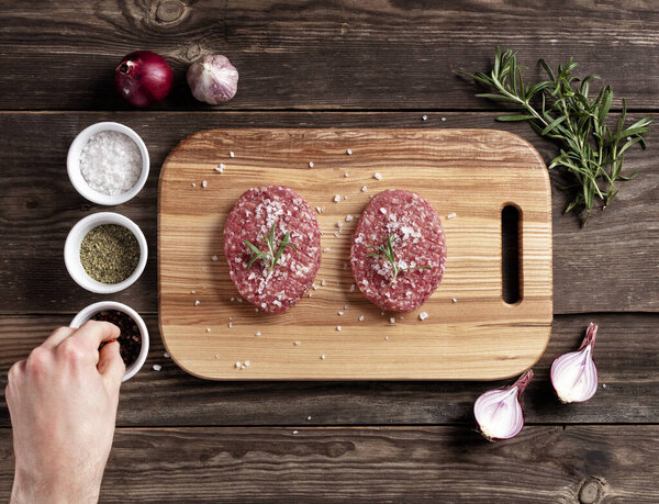 Raw meat round cutlets - for cooking homemade hamburgers. Cutlets on a wooden cutting board. The cook takes pepper for meat seasoning. View from above. Flat lay.
