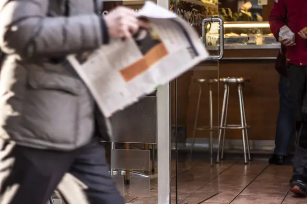 A man reading a newspaper walking down the street of Barcelona. In the background is a small coffee shop with pastries. The man is out of focus. The main focus on the routine of urban turbulent life.