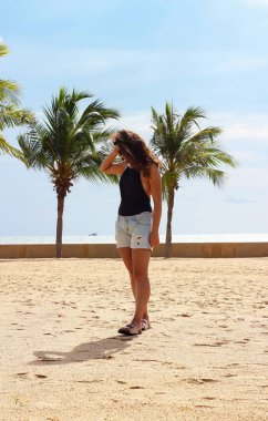 Kumlu sahilde, okyanus kıyısında palmiye ağaçları olan güzel, ince bir kız. Tatil kavramı.
