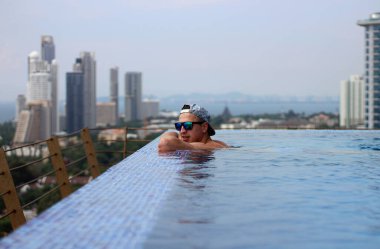 Şapkalı ve gözlüklü gülümseyen genç adam gökdelendeki havuzun manzarasının keyfini çıkarıyor..