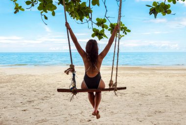 Mayolu genç ve çekici bir kız okyanusun kıyısındaki kumlu bir sahilde salıncakta oturuyor ve manzaranın tadını çıkarıyor. Tatil kavramı.