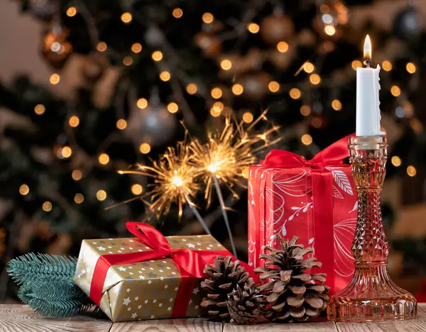 Christmas composition with burning candle, gifts and cones on the background of a decorated Christmas tree and burning sparklers.