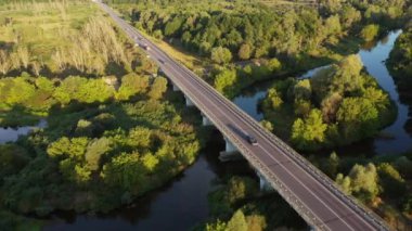 Gündoğumunda güzel bir sonbahar ormanında nehrin üzerinde köprüsü olan bir otoyol. Köprüden geçen arabaların hava görüntüsü.