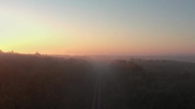Güneşin doğuşunda demiryolu olan sisli bir sonbahar ormanının havadan görünüşü ve arka planda şehrin silueti..