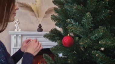 Noel arifesinde ağacı süslemeye başlayın. Elinde altın bir küre tutan genç bir kadın onu yanan şöminenin önündeki köknar dalına asıyor. Kış tatili atmosferi
