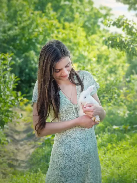 Yeşil bahçe arka planında beyaz bir tavşanı kollarında tutan genç bir kadının şirin portresi. Sevimli bir tavşanı okşayan kız