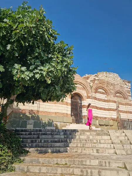 Bulgaristan 'ın Nessebar kentindeki eski tiyatronun yakınlarındaki St. John Aliturgetos Ortaçağ Kilisesi' nin üst merdivenlerinden manzarayı izleyen pembe elbiseli hoş bir genç kadın.