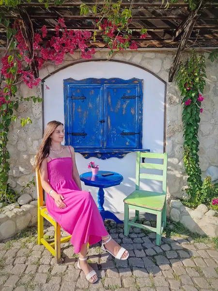 Kafe terasında oturan pembe elbiseli estetik genç kadın güneşli günün tadını çıkarıyor. Restoranda renkli bir açık hava sahnesi. Sarı ve yeşil sandalyelerle çevrili mavi masa, arka planda mavi pencere.