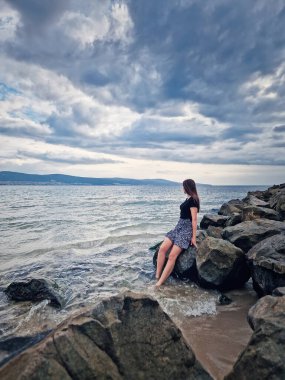 Deniz kenarındaki kayalarda oturan genç bir kadın bulutlu bir gökyüzünün altında ufka doğru bakıyor. Huzurlu manzara, engin okyanus ve gökyüzünün sakin bir atmosfer yaratmasıyla derin bir tefekkür hissi uyandırıyor.