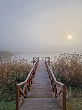 Göl kıyısındaki ahşap iskele sisli bir sahnenin içine açılıyor, uzun kuru kamışlarla çevrili. Güneşin doğuşu sislerin arasından belli belirsiz görülür, sakin ve sakin bir atmosfer yaratır. Sakin ve ruhani bir sahne.