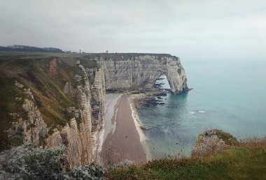 Fransa 'nın Normandiya kentindeki ünlü Etretat uçurumları manzaralı, çakıl taşı plajı, turkuaz sular ve bulutlu gökyüzü altındaki yemyeşil tepeler yakınlarında doğal Porte d' Aval kemer oluşumuna ev sahipliği yapıyor.