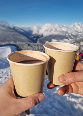 Two friends hands holding paper cups filled with hot chocolate beverage on the top of the a snowy mountain. Winter activity and vacation enjoyment concept clipart