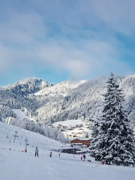 La Clusaz, Annecy, Fransa 'da yamaçları, asansörleri ve kayakçıları olan dağ kayak merkezi manzarası. Ağaçlar karla kaplı ve vadide kulübeleri olan küçük bir köy.