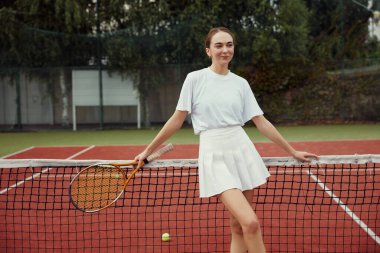 Tenis raketiyle korttaki tenis sahasında poz veren beyaz elbiseli şık bir kadın. Spor Moda.
