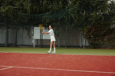 Beyazlar içinde tenis oyuncusu bir maçta topu servis ediyor. Mahkemede tenis oynayan genç bir kız..