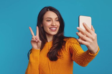 Sıcak örülmüş turuncu süveter giyen neşeli güzel kız mavi arka planda akıllı telefonuyla selfie çekerken vsign hareketi yapıyor.