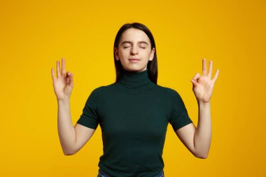 Sakin, rahat görünümlü esmer kız stres, kapalı gözler ve huzur içinde meditasyon yaparken gülümsüyor. Zen, ayakta nilüfer duruşu, yoga, sarı arka plan.